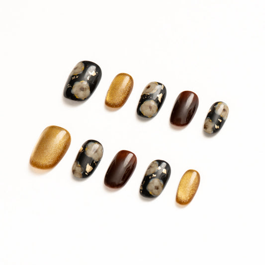 Flat lay of ten press-on nails arranged in two rows on a white background, featuring a black, gold, and deep brown color palette. The set includes glossy black nails with soft floral motifs and scattered gold foil, rich chocolate-brown solids, and bold metallic gold shimmer nails for an elegant, high-contrast statement look.