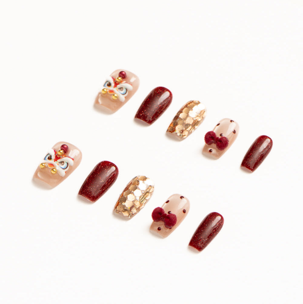 Flat lay of ten press-on nails arranged in two rows on a white background, featuring a festive red, gold, and nude color palette. The set includes glossy deep red shimmer nails, sheer nude accents with red polka dots and plush velvet bow details, gold foil mosaic textures, and statement nails with intricate mask-inspired designs accented by pearls and metallic beads for a bold, celebratory look. Lunar New Year nails
