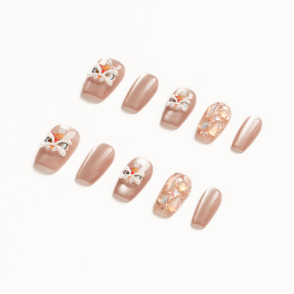 Flat lay of ten press-on nails arranged in two rows on a white background, featuring a soft nude-beige palette. The set includes glossy solid nude nails, accent nails with delicate pearl details, abstract iridescent shell textures, and playful hand-painted mask-inspired designs with white, red, and gold accents for an elegant yet whimsical look. Lunar New year nails