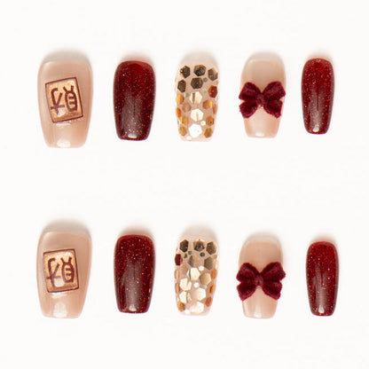 Flat lay of ten press-on nails arranged in two rows on a white background, featuring a deep burgundy and nude color palette. The set includes glossy wine-red shimmer nails, sheer nude accents with gold foil mosaic texture, plush velvet bow embellishments, and a minimalist nude nail with a small gold square charm for a luxe, lunarnewyear-inspired look.