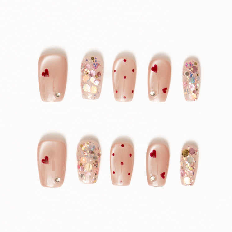 Flat lay of ten press-on nails arranged in two rows on a white background, featuring a sheer nude base with playful red heart accents, delicate red polka dots, and shimmering confetti glitter details. The design blends soft romance with subtle sparkle for a charming, celebratory look.