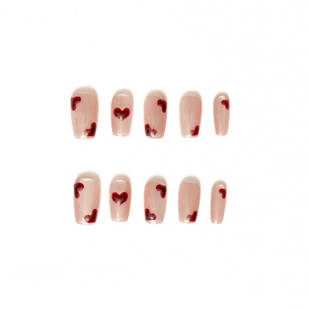 Flat lay of ten press-on nails arranged in two rows on a white background, featuring a sheer nude-pink base with scattered glossy red heart accents. The minimalist, romantic design has a soft, delicate finish perfect for a sweet, modern Valentine-inspired look.