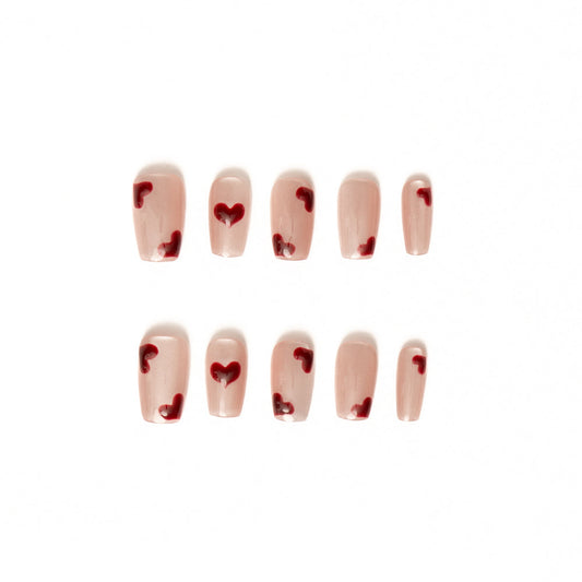 Flat lay of ten press-on nails arranged in two rows on a white background, featuring a sheer nude-pink base with scattered glossy red heart accents. The minimalist, romantic design has a soft, delicate finish perfect for a sweet, modern Valentine-inspired look.