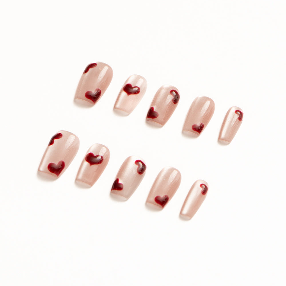 Flat lay of ten press-on nails arranged in two rows on a white background, featuring a sheer nude-pink base with scattered glossy red heart accents. The minimalist, romantic design has a soft, delicate finish perfect for a sweet, modern Valentine-inspired look.