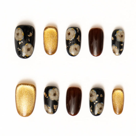 Flat lay of ten press-on nails arranged in two rows on a white background, featuring a black, gold, and deep brown color palette. The set includes glossy black nails with soft floral motifs and scattered gold foil, rich chocolate-brown solids, and bold metallic gold shimmer nails for an elegant, high-contrast statement look.