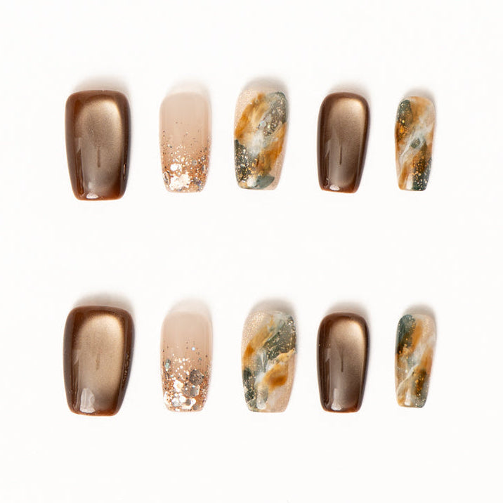 Flat lay of ten press-on nails arranged in two rows on a white background, featuring a warm bronze and earth-tone color palette. The set includes glossy metallic bronze nails, sheer nude accents with scattered gold flecks, and abstract marble designs blending moss green, amber, cream, and gold for an organic, stone-inspired look.