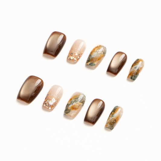 Flat lay of ten press-on nails arranged in two rows on a white background, featuring a warm bronze and earth-tone color palette. The set includes glossy metallic bronze nails, sheer nude accents with scattered gold flecks, and abstract marble designs blending moss green, amber, cream, and gold for an organic, stone-inspired look.