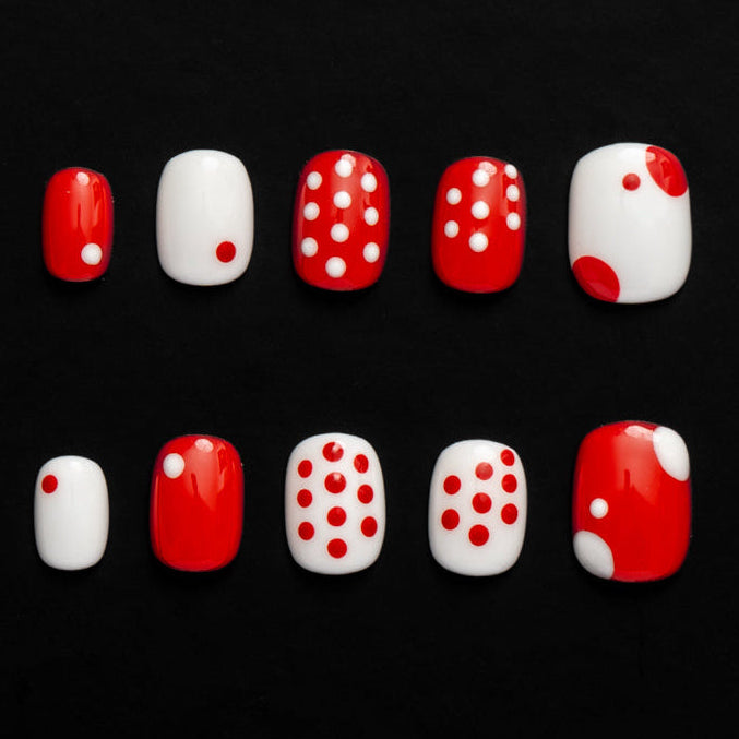 Flat lay of ten press-on nails arranged in two rows on a black background, featuring a bold red and white color palette. The set includes glossy solid red and white nails, playful polka-dot designs, and contrasting accent patterns for a clean, graphic, and retro-inspired look.